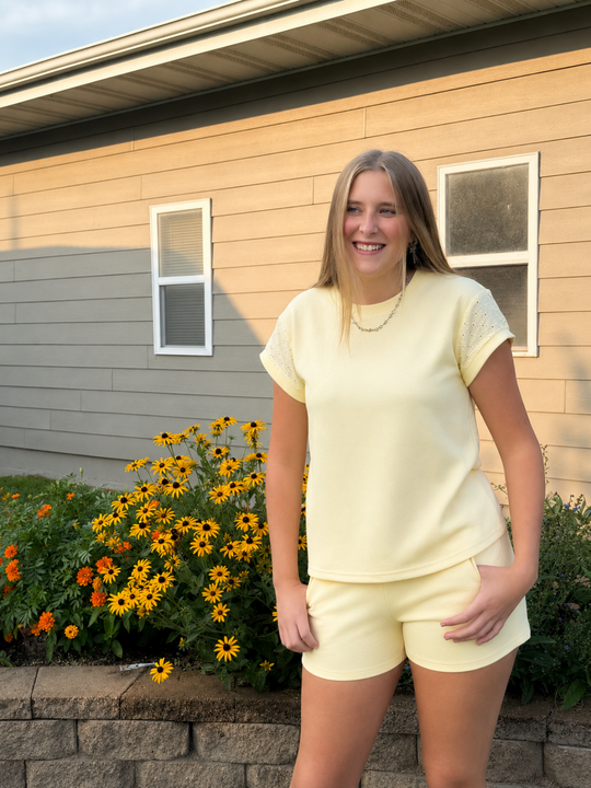 A woman stands outdoors in front of a house, wearing a pale yellow short-sleeve top and matching shorts. Yellow and orange flowers are visible in the garden bed behind her.