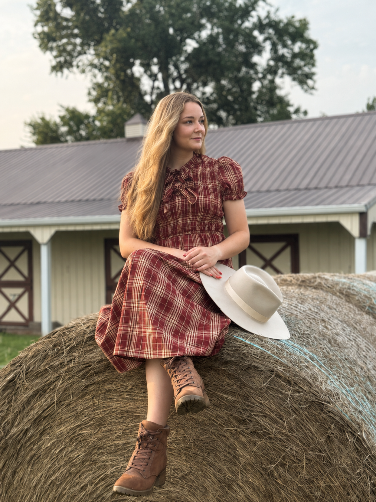 A young woman with long blonde hair sits on a large hay bale in front of a barn, wearing a red and brown plaid short-sleeve dress, brown lace-up boots, and holding a wide-brimmed cream hat.